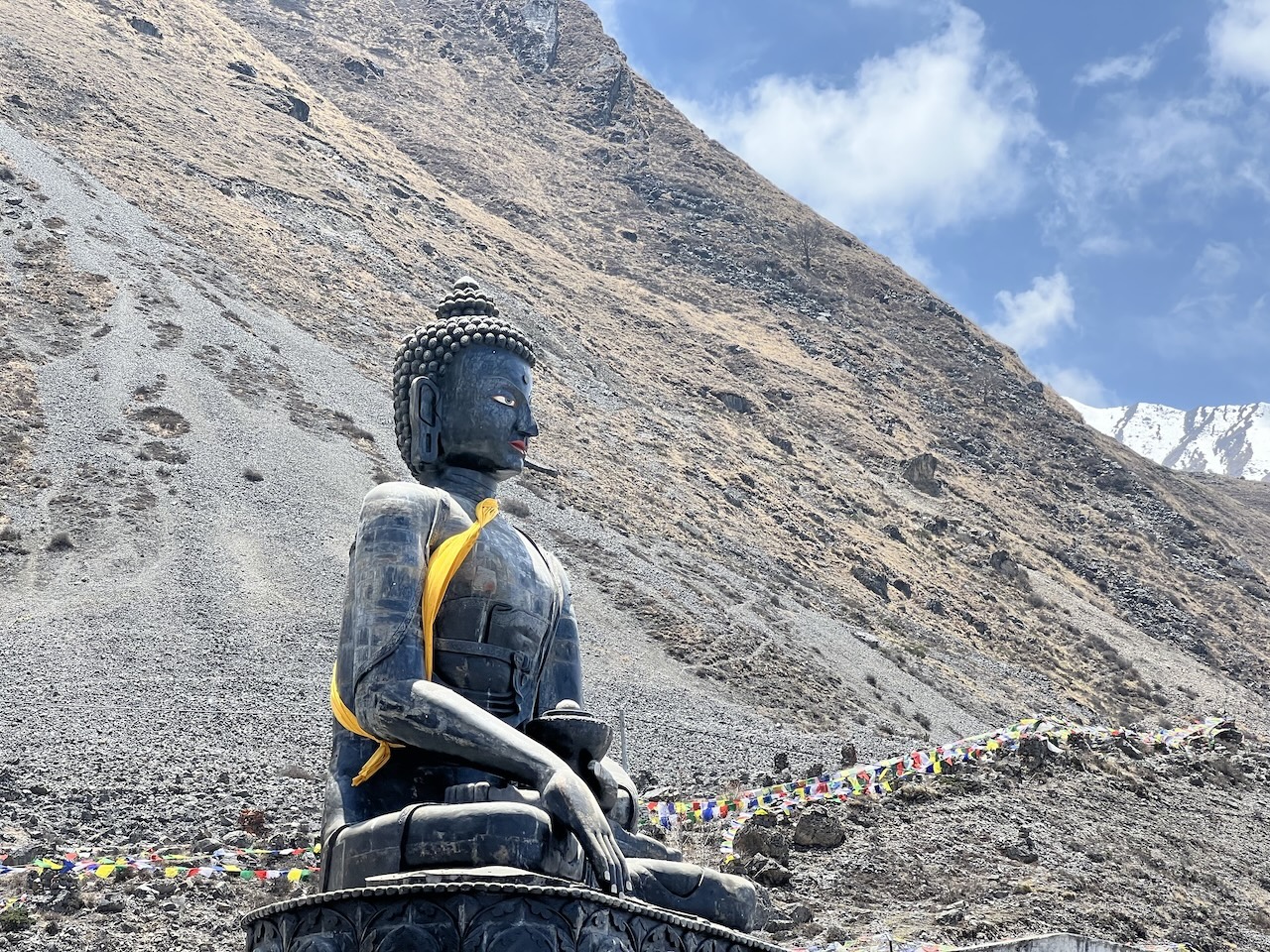 Statua del Buddha seduto con drappo giallo, bandiere di preghiera e pendii rocciosi sullo sfondo