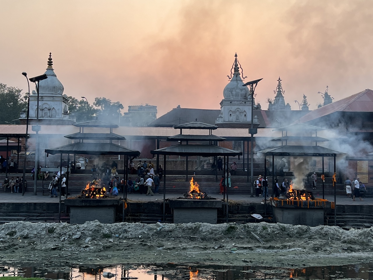 Roghi funerari su bracieri lungo il fiume, fumo denso e padiglioni con templi bianchi sullo sfondo.