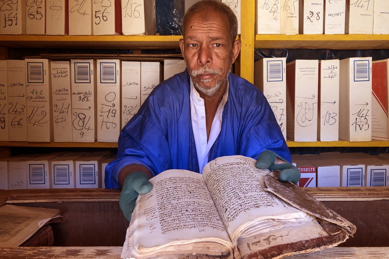 Un bibliotecario mauritano di Chinguetti mostra un manoscritto antico, circondato da scaffali di libri.