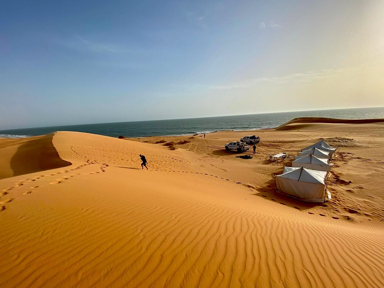 Tende bianche per accampamento turistico nel deserto, con dune di sabbia dorata, oceano sullo sfondo e cielo azzurro.