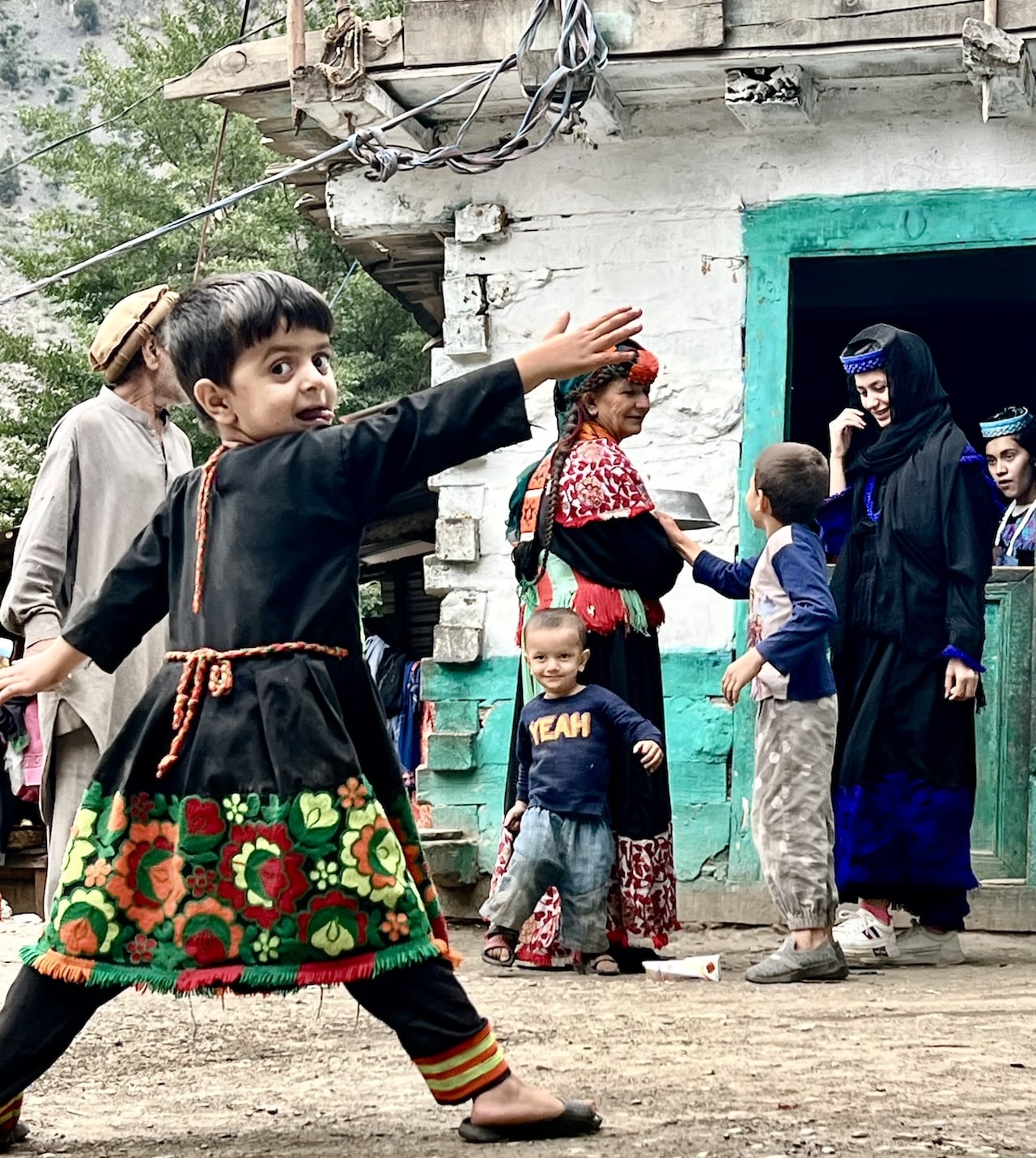 Bambini Kalash in abiti tradizionali decorati che giocano all'aperto davanti a una casa, adulti sullo sfondo