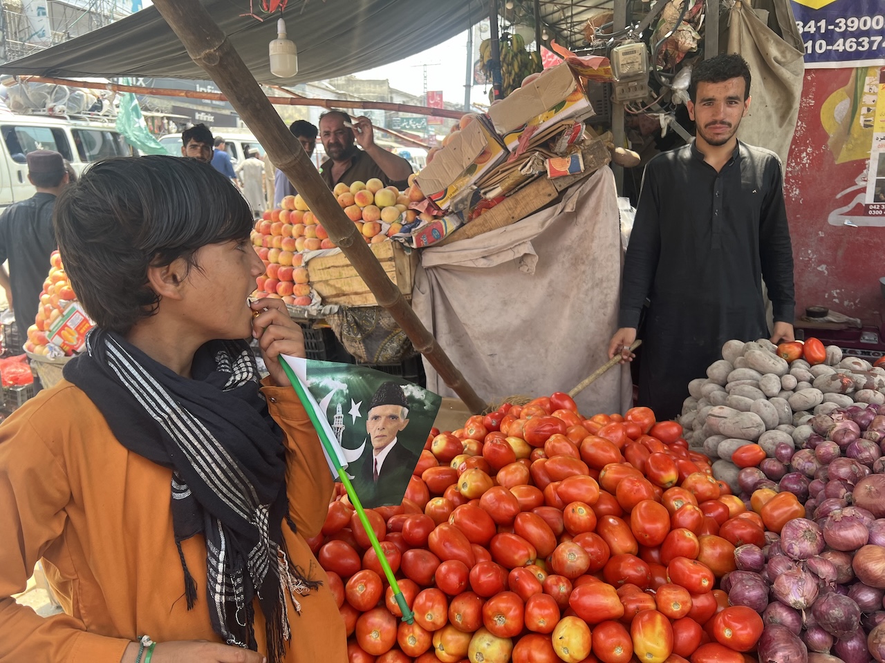 Bancarella di frutta e verdura, con un ragazzo che tiene una bandiera pakistana in mezzo a mucchi di pomodori e cipolle