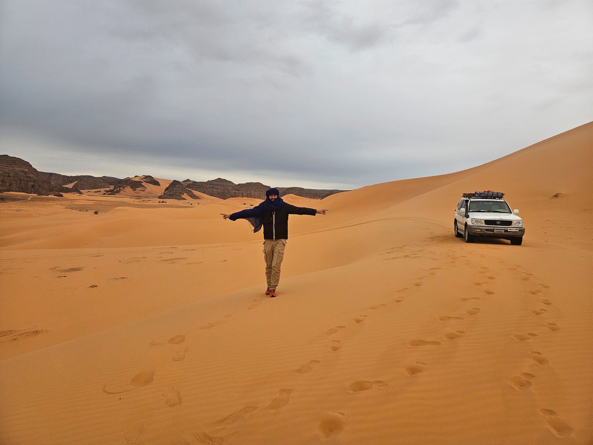 Viaggiatore con braccia aperte sulle dune del deserto algerino, fuoristrada sullo sfondo durante il tour