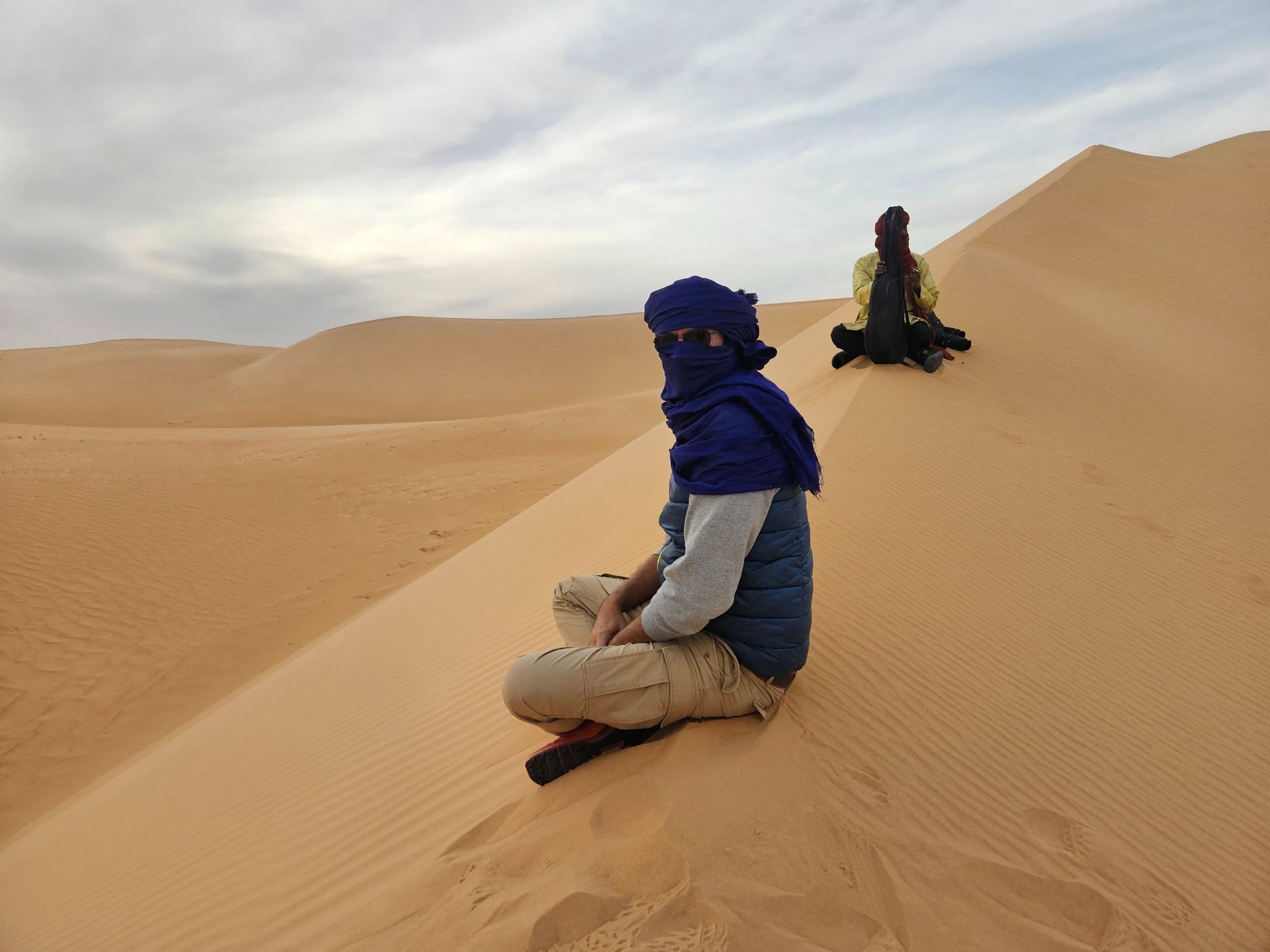 Viaggiatore con turbante blu tuareg seduto sulla duna, sullo sfondo la guida con la chitarra tradizionale