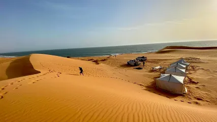 Tende bianche per accampamento turistico nel deserto, con dune di sabbia dorata, oceano sullo sfondo e cielo azzurro.