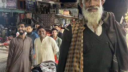 Uomo anziano barbuto in cappello e gilet posa di notte per strada a Naran. Altri uomini sorridenti dietro di lui.