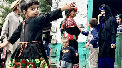 Bambini Kalash in abiti tradizionali decorati che giocano all'aperto davanti a una casa, adulti sullo sfondo